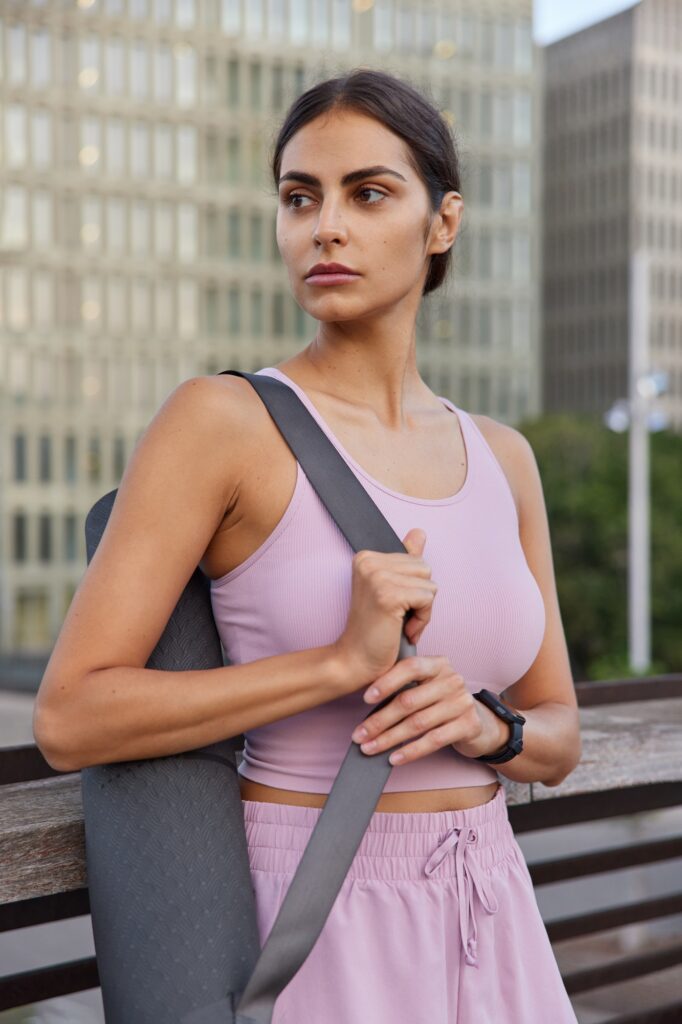 Outdoor shot of pensive sportswoman wears tank top and shorts carries yoga mat prepares for pilates